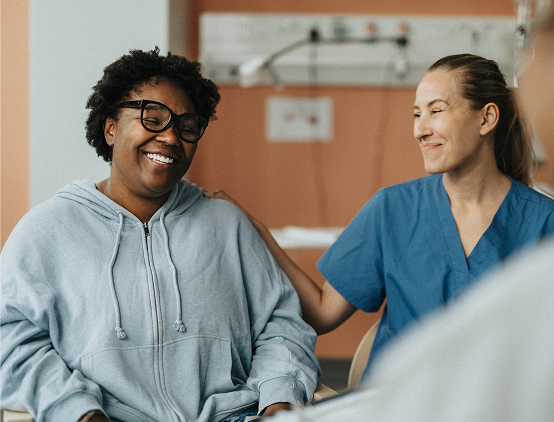 Woman in gray sweater smiling while comforted by woman in blue scrubs